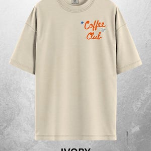 Może przedstawiać: Kremowa koszulka z kr&oacute;tkim rękawem. Koszulka ma napis "Coffee Club" pomarańczową czcionką z niebieską gwiazdką i słowami "Limited Time Only" w kolorze niebieskim. Słowo "IVORY" jest wydrukowane pogrubioną, białą czcionką na dole.