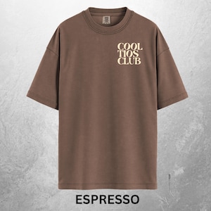 May include: Espresso-colored t-shirt with the text "COOL TIOS CLUB" in a cream-colored font on the upper chest. The shirt has short sleeves and a crew neck. The word "ESPRESSO" is printed at the bottom of the shirt.
