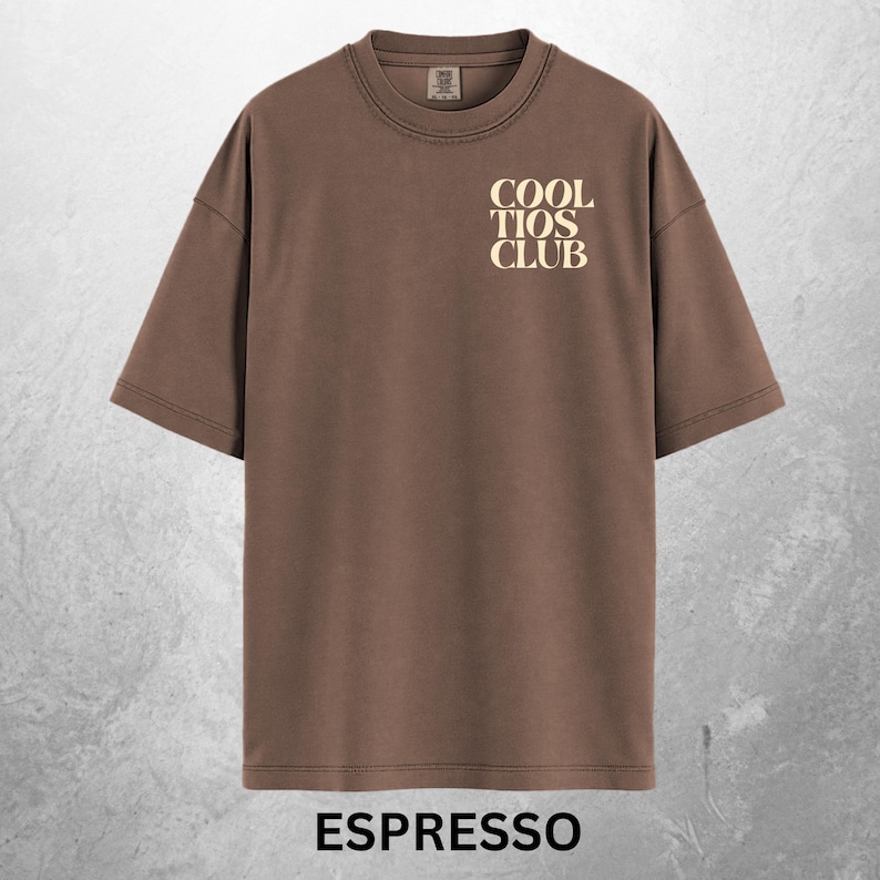 May include: Espresso-colored t-shirt with the text "COOL TIOS CLUB" in a cream-colored font on the upper chest. The shirt has short sleeves and a crew neck. The word "ESPRESSO" is printed at the bottom of the shirt.