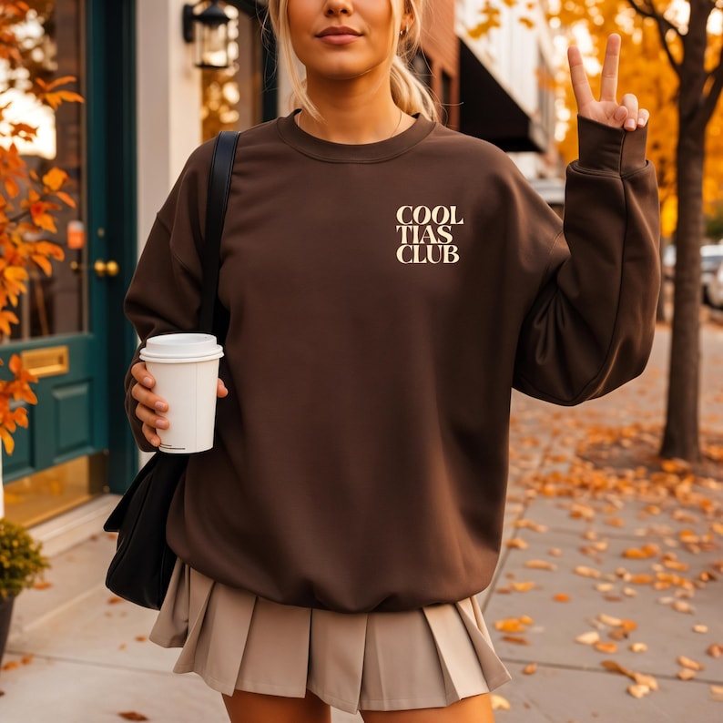 May include: A brown sweatshirt with the text "COOL TIAS CLUB" in a cream-colored font. The sweatshirt is paired with a beige pleated skirt. The person is holding a white coffee cup and a black shoulder bag.