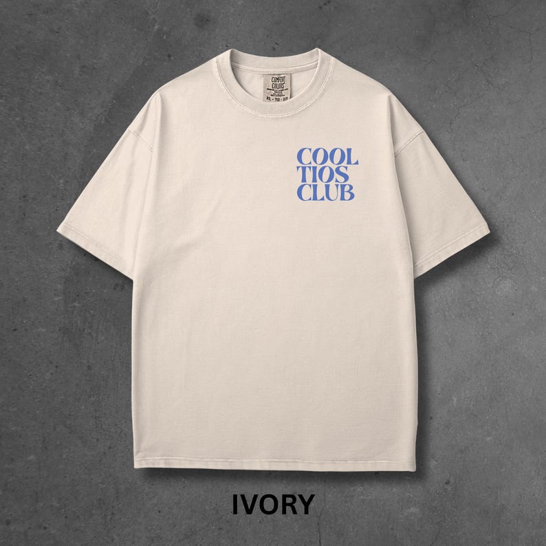 May include: Ivory-colored t-shirt with the text "COOL TIOS CLUB" in blue. The shirt has a crew neck and short sleeves. The word "IVORY" is printed at the bottom of the shirt.