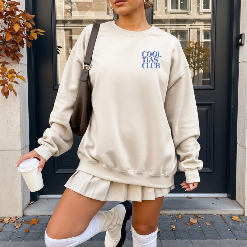 May include: A beige sweatshirt with the text "COOL TIAS CLUB" in blue. The sweatshirt is paired with a pleated skirt, white knee-high socks, and white boots. A brown shoulder bag and a white coffee cup complete the outfit.