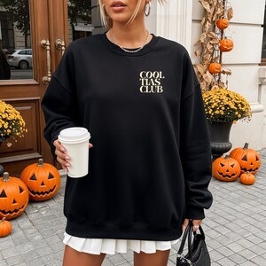 May include: Black sweatshirt with the text "COOL TIAS CLUB" in a gold color. The sweatshirt is paired with a white pleated skirt. The background includes Halloween decorations, such as pumpkins and fall foliage.