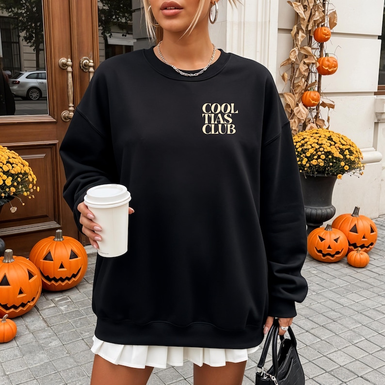 May include: Black sweatshirt with the text "COOL TIAS CLUB" in a gold color. The sweatshirt is paired with a white pleated skirt. The background includes Halloween decorations, such as pumpkins and fall foliage.