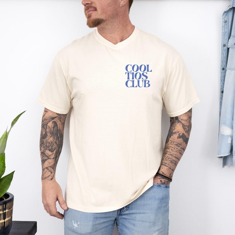 May include: Cream-colored t-shirt with the text "COOL TIOS CLUB" in blue. The shirt has a classic crew neck and short sleeves. The text is centered on the chest. The model is wearing blue jeans.