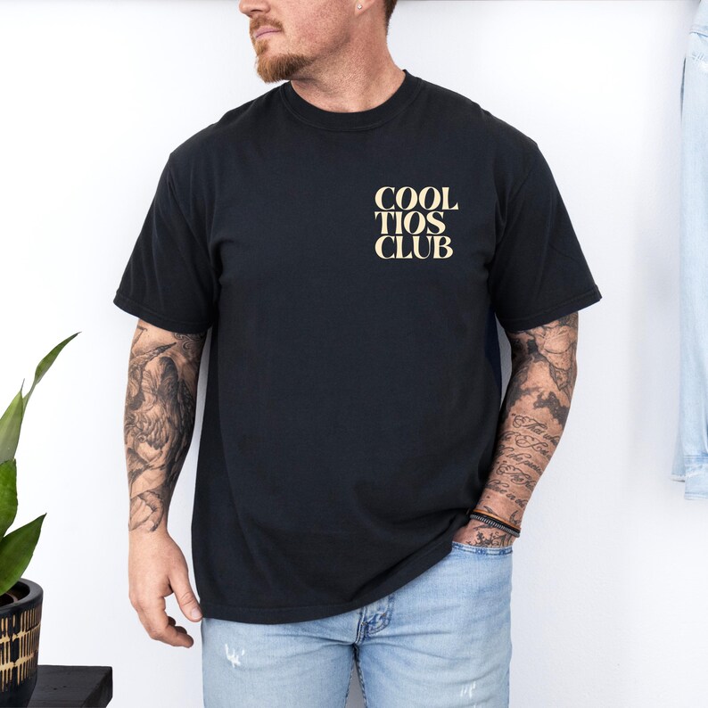 May include: Black t-shirt with the text "COOL TIOS CLUB" in a light tan color on the upper left chest. The shirt is a classic crew neck style and appears to be made of a soft material.
