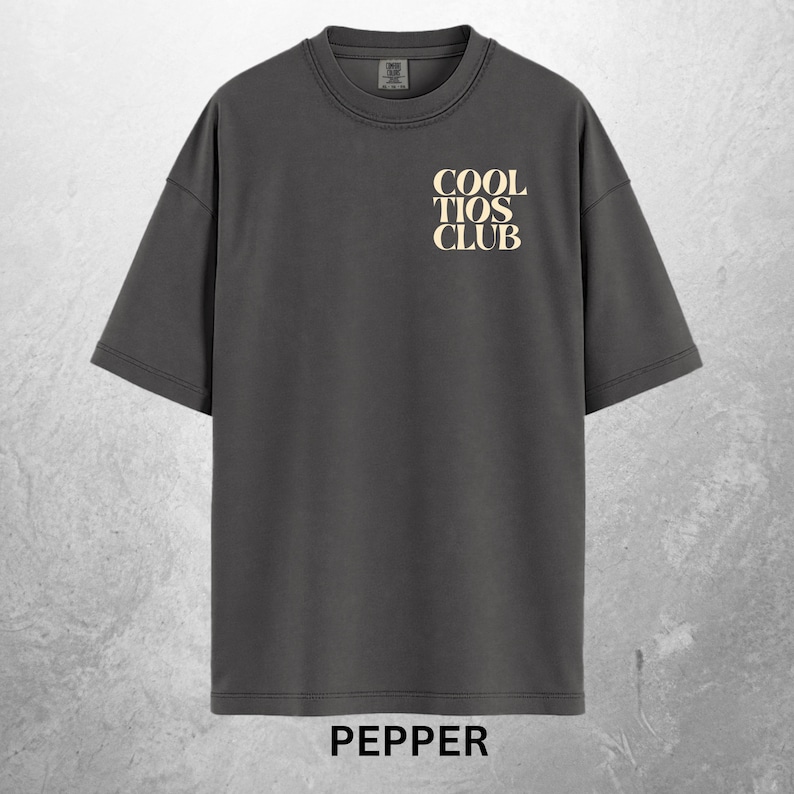 May include: A dark gray t-shirt with the text "COOL TIOS CLUB" in a cream color on the upper chest. The word "PEPPER" is printed at the bottom of the shirt. The shirt has short sleeves and a crew neck.