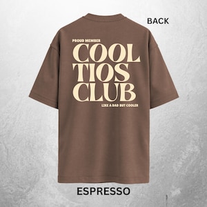 May include: Espresso-colored t-shirt with the text "COOL TIOS CLUB" in a bold, cream-colored font. The shirt also features the phrases "PROUD MEMBER" and "LIKE A DAD BUT COOLER". The word "BACK" is printed at the top of the shirt.