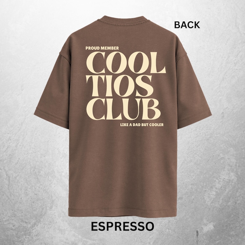 May include: Espresso-colored t-shirt with the text "COOL TIOS CLUB" in a bold, cream-colored font. The shirt also features the phrases "PROUD MEMBER" and "LIKE A DAD BUT COOLER". The word "BACK" is printed at the top of the shirt.