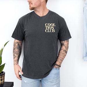 May include: Dark gray t-shirt with a V-neck. The shirt features the text "COOL TIOS CLUB" in a light tan color on the upper left chest. The shirt is worn by a person wearing blue jeans.