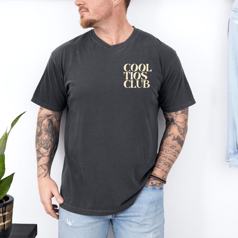 May include: Dark gray t-shirt with a V-neck. The shirt features the text "COOL TIOS CLUB" in a light tan color on the upper left chest. The shirt is worn by a person wearing blue jeans.
