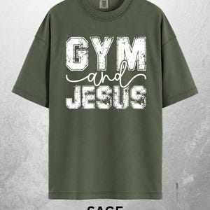 May include: Sage green t-shirt with the words "GYM and JESUS" in distressed white lettering. The shirt has a crew neck and short sleeves. The word "SAGE" is printed at the bottom of the shirt.