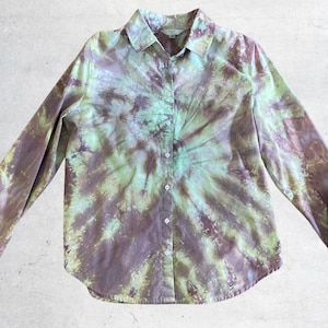 May include: A long-sleeve button-down shirt featuring a spiral tie-dye pattern in shades of muted purple and light lime green. The garment has a standard collar, a full-length front button placket, and a curved hemline at the bottom edge.