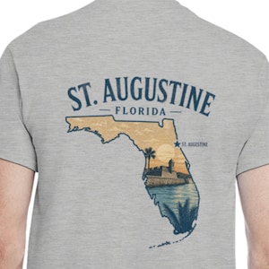 May include: A light gray t-shirt featuring a graphic of Florida with a sunset scene and the text "ST. AUGUSTINE FLORIDA". The design includes a palm tree, a fort, and the text "ST. AUGUSTINE".