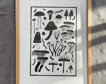 May include: A framed print featuring a black and white linocut illustration of various mushroom species. The artwork is set against a white background and framed in a natural wood frame. The print is a botanical theme.