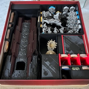 May include: A red and black game box with a custom insert for organising game pieces. The insert has compartments for cards, miniatures, and other game components. The box is open and the insert is visible.