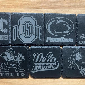 May include: Set of 8 black slate coasters with different college logos and mascots. The logos include USC Trojans, Ohio State, Penn State, Oregon Ducks, Notre Dame Fighting Irish, UCLA Bruins, and the University of Michigan Wolverines.