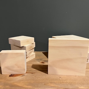 Puede incluir: Una pila de seis cuadrados de madera sin terminar, cada uno mide aproximadamente 10 cm por 10 cm, y un solo cuadrado de madera sin terminar más grande que mide aproximadamente 15 cm por 15 cm. Los cuadrados son todos de madera de color claro y tienen una superficie lisa.