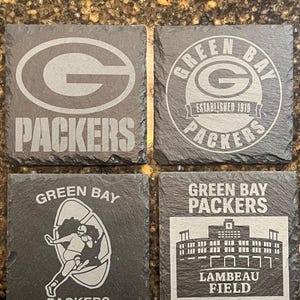 May include: Four square slate coasters featuring the Green Bay Packers logo and imagery. Designs include the team's "G" logo, a circular logo, a football player graphic, and Lambeau Field. The coasters are dark gray with white graphics.