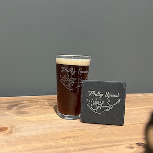 May include: A glass of dark brown beer with a white foam head in a clear pint glass with the text "Philly Special" and a football play diagram. A black slate coaster with the same text and diagram is next to the glass.