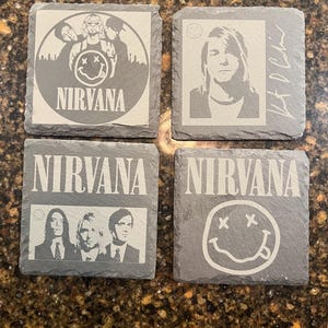 May include: Set of four black slate coasters featuring the band Nirvana. Each coaster has a different image, including a band photo, a smiley face logo, and a solo portrait of Kurt Cobain. The text "Nirvana" is printed on two of the coasters.