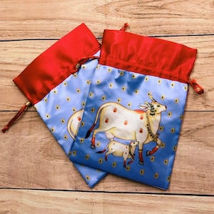 May include: Two blue drawstring bags with red trim. The bags feature a white cow and calf with gold accents on a blue background with small yellow dots.