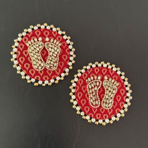 May include: Two round, red fabric decorations with a repeating pattern. Each has a beaded border of white and gold beads. The center of each decoration features a pair of bejeweled footprints, with clear, sparkling stones.