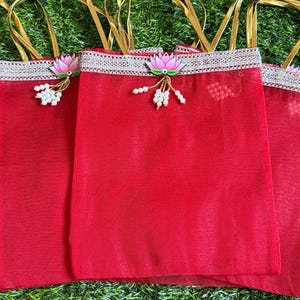 May include: Three red sheer fabric gift bags with gold ribbon handles. Each bag features a silver metallic trim along the top edge, decorated with a pink lotus flower emblem and a cluster of small white beads hanging from the center of the flower.