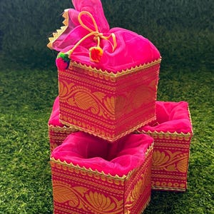 May include: Three square containers featuring a vibrant hot pink fabric base with intricate gold metallic floral embroidery. Each container is trimmed with gold scalloped edging and includes a matching pink drawstring pouch top with colorful pom-poms.