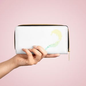 Puede incluir: Cartera blanca con cremallera y un diseño abstracto colorido de una ola con estrellas. La cartera está en una mano.