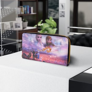 Puede incluir: Una cartera con cremallera de color morado y azul con una imagen impresa de los personajes de la película Frozen II. La imagen incluye a Elsa, Anna, Kristoff, Sven y Olaf de pie en un paisaje nevado.
