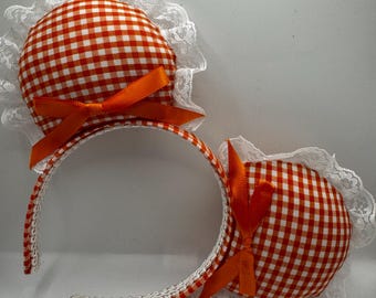 May include: A headband featuring two round, red and white gingham fabric ears trimmed with white lace. Each ear has an orange satin bow. The headband itself is covered in matching gingham fabric and white lace.