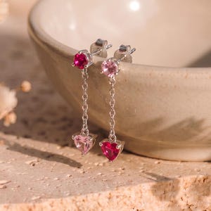 May include: A pair of silver drop earrings featuring a small round pink or red gemstone at the top, connected by a delicate silver chain to a heart-shaped gemstone pendant. One earring has a dark pink heart, while the other has a light pink heart.