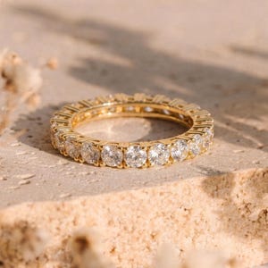 May include: A gold eternity ring with a continuous row of round-cut, clear gemstones. The ring is set on a light-colored, textured surface, with soft lighting and a blurred background. The ring's band is a bright, polished gold.