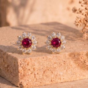 May include: A pair of gold-colored stud earrings. Each earring features a central, round, deep red gemstone surrounded by a halo of smaller, clear gemstones. The earrings are set on a textured, light brown surface.