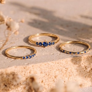May include: Three gold rings, each set with blue gemstones, are arranged on a textured, light-toned surface. The rings showcase different designs: a row of small stones, a cluster, and evenly spaced gems, offering a variety of styles.