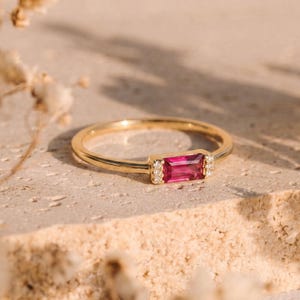 May include: A gold ring with a rectangular, magenta-colored gemstone at the center. Small, clear stones flank the main gem. The ring is set on a light-colored, textured surface, with soft, blurred floral elements in the background.