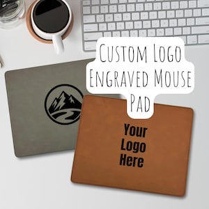 May include: Two custom engraved mouse pads. One is gray with a mountain and river logo, the other is brown with the text "Your Logo Here". The text "CUSTOM LOGO ENGRAVED MOUSE PAD" is also visible.