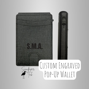 May include: A black custom engraved pop-up wallet with the initials "S.M.A." printed on the front. The wallet is shown from the front and side, highlighting its slim design. The Sandpiper Ink logo is visible in the lower left corner.