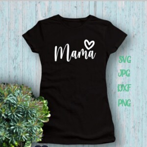 May include: Black t-shirt with the word "Mama" in white script and a small heart. The shirt is displayed on a light blue and white wooden background. A small plant in a green pot is in the lower left corner.