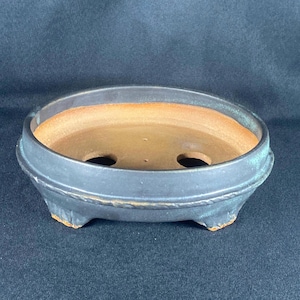 May include: A black ceramic bonsai pot with a round shape and three small feet. The interior is a light brown colour, and the pot has two drainage holes. The rim has a decorative ridge.