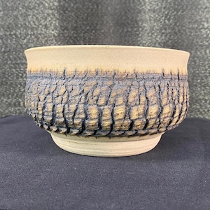 Peut inclure: Un bol en céramique fait à la main avec un motif texturé en forme d'écorce dans des tons beige, bronzé et bleu foncé. Le bol a une forme arrondie avec un bord légèrement évasé et une base solide. Cette pièce unique est idéale pour servir ou exposer.