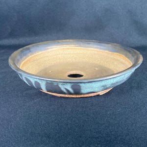 Peut inclure: Un pot à bonsaï rond en céramique avec un bord et un extérieur bleu foncé. L'intérieur est de couleur beige clair. Le pot a un trou de drainage au centre et est conçu pour planter de petits arbres ou plantes.