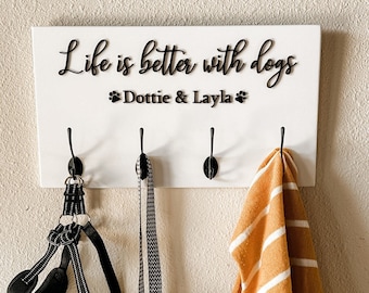 May include: White wooden coat rack with the text "Life is better with dogs" and the names "Dottie & Layla" with paw prints. The rack has four hooks with a black dog leash, a black dog harness, and a yellow and white striped sweater hanging on them.