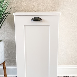 May include: White wooden cabinet with a single door and a black metal handle. The cabinet is standing on a black and white patterned rug.