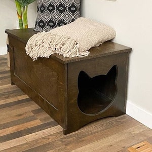 May include: A dark brown wooden storage bench featuring a cat-shaped cutout entrance on the front panel. A black and white geometric patterned throw pillow and a beige knit throw blanket with fringe edges are placed on top of the bench surface.