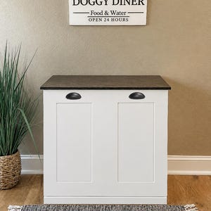 May include: A white cabinet with a dark brown top and two black cup pulls. Above the cabinet is a rectangular white sign with black text that reads: Ernie's DOGGY DINER, Food & Water, OPEN 24 HOURS, featuring paw print icons on the sides.
