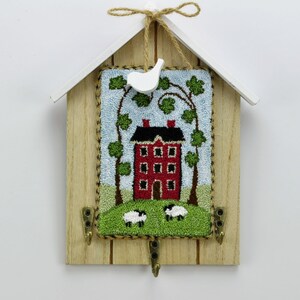 Op de afbeelding: Een houten sleutelhanger in de vorm van een huis met een rood huis en groene bomen aan de voorkant. Er zijn twee haken aan de onderkant om sleutels aan te hangen.