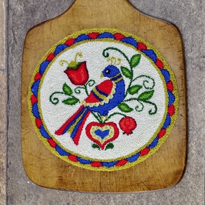 May include: A wooden cutting board with a hand-stitched design of a blue bird with red and yellow accents. The design is surrounded by a blue, red, and gold border.
