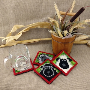 May include: Four square-shaped coasters with a green background and red border. Each coaster features a different sheep design with black and white wool. The coasters are on a brown burlap surface.